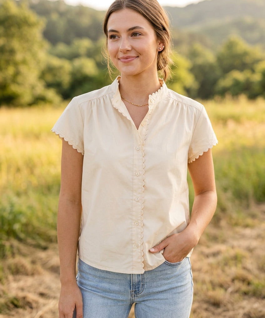 A cream-colored short-sleeve blouse with a ruffled mock neck and button-front detailing, displayed on a dress form against a light blue paneled wall.