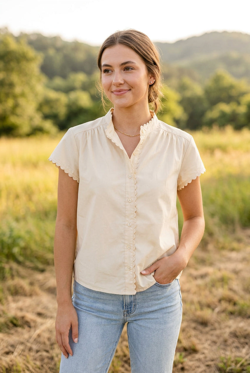 A cream-colored short-sleeve blouse with a ruffled mock neck and button-front detailing, displayed on a dress form against a light blue paneled wall.